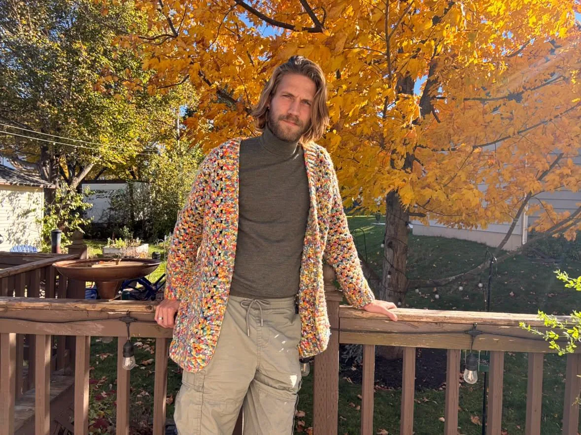 Vic leaning on a wooden deck rail under a golden autumn maple, wearing a speckled multicoloured crocheted cardigan