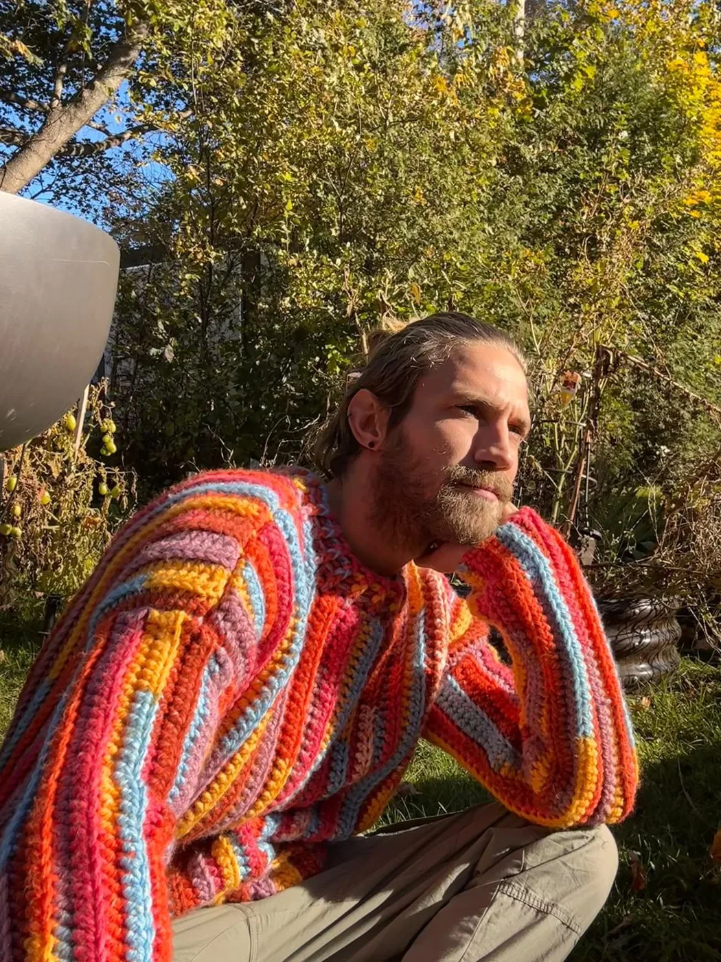 Vic sitting in a garden in a chunky crocheted jumper striped in red, orange, yellow and blue