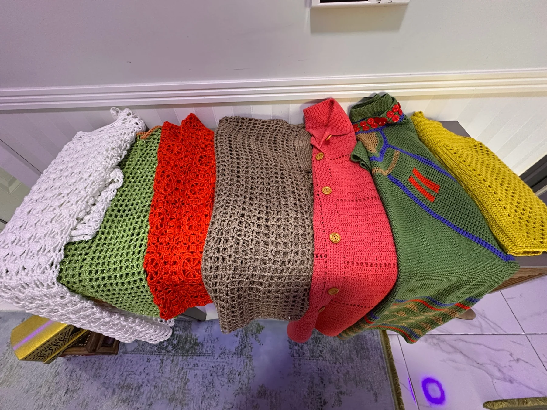 Seven crocheted garments folded side by side, from white lace to coral, green and mustard yellow
