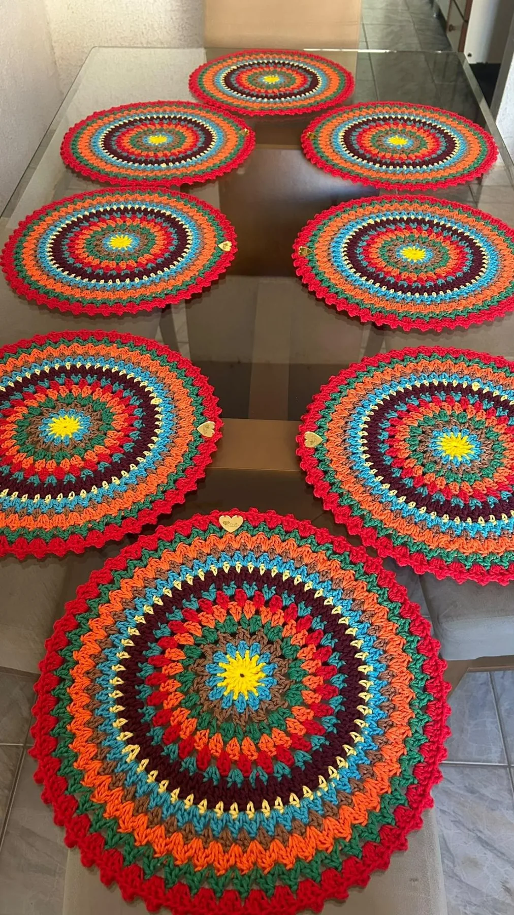 Nine round crocheted mandalas in red, orange, blue, green and yellow, laid out on a glass table