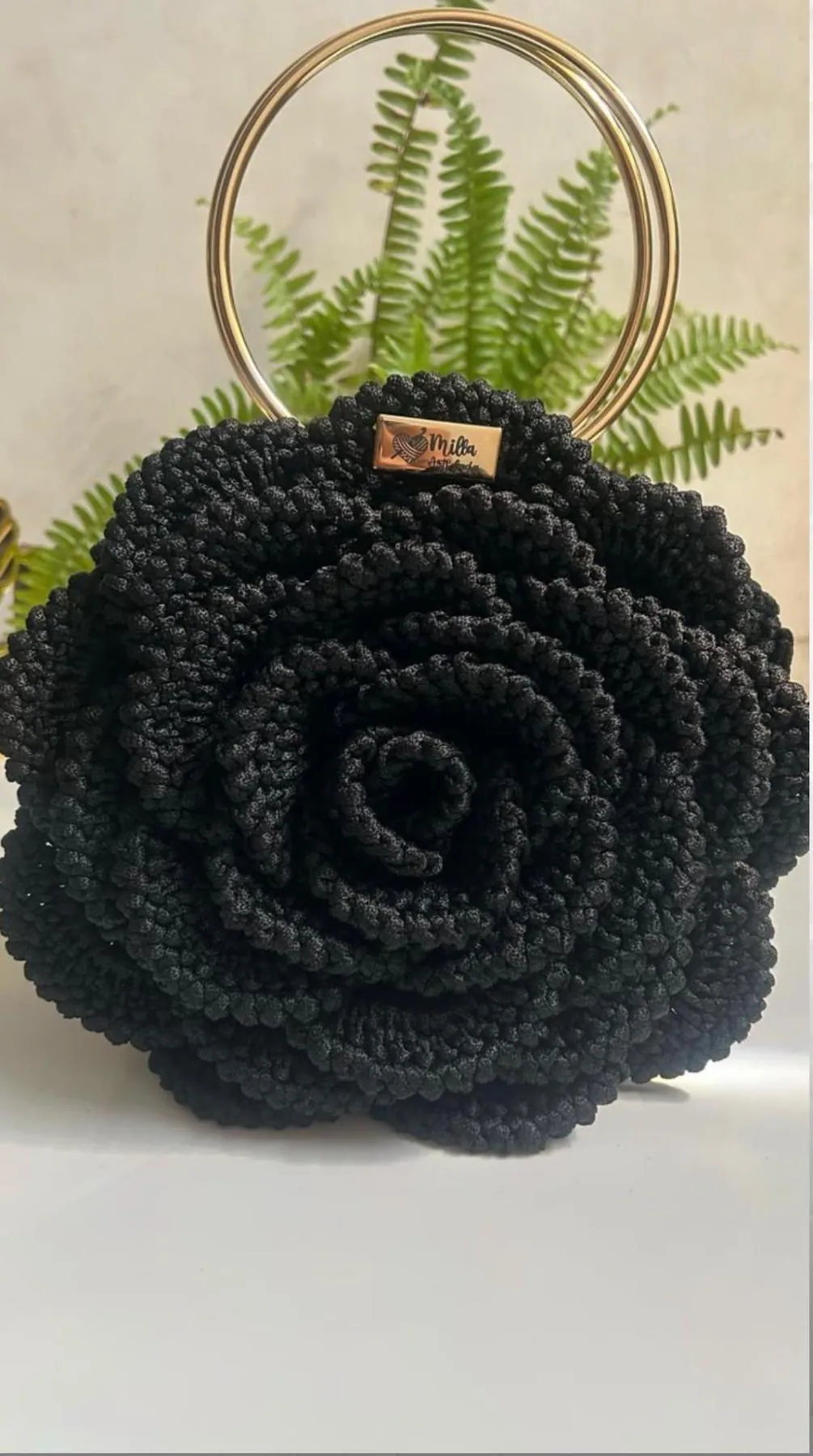 Black crocheted bag shaped like a large flower, with a round golden handle and a small Milla Arts Crochês label
